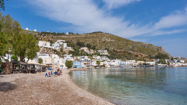 Un pueblo costero con edificios en una ladera, una playa de guijarros y aguas tranquilas.