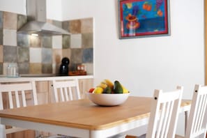 A dining area with a white table and chairs, a bowl of fruit on the table, a kitchen in the background, and a colorful painting on the wall.