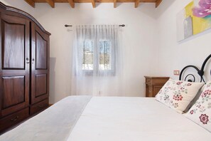 Un dormitorio con un armario de madera, una cama con almohadillas estampadas, un ventanal con cortinas translúcidas y un cabecero de madera.