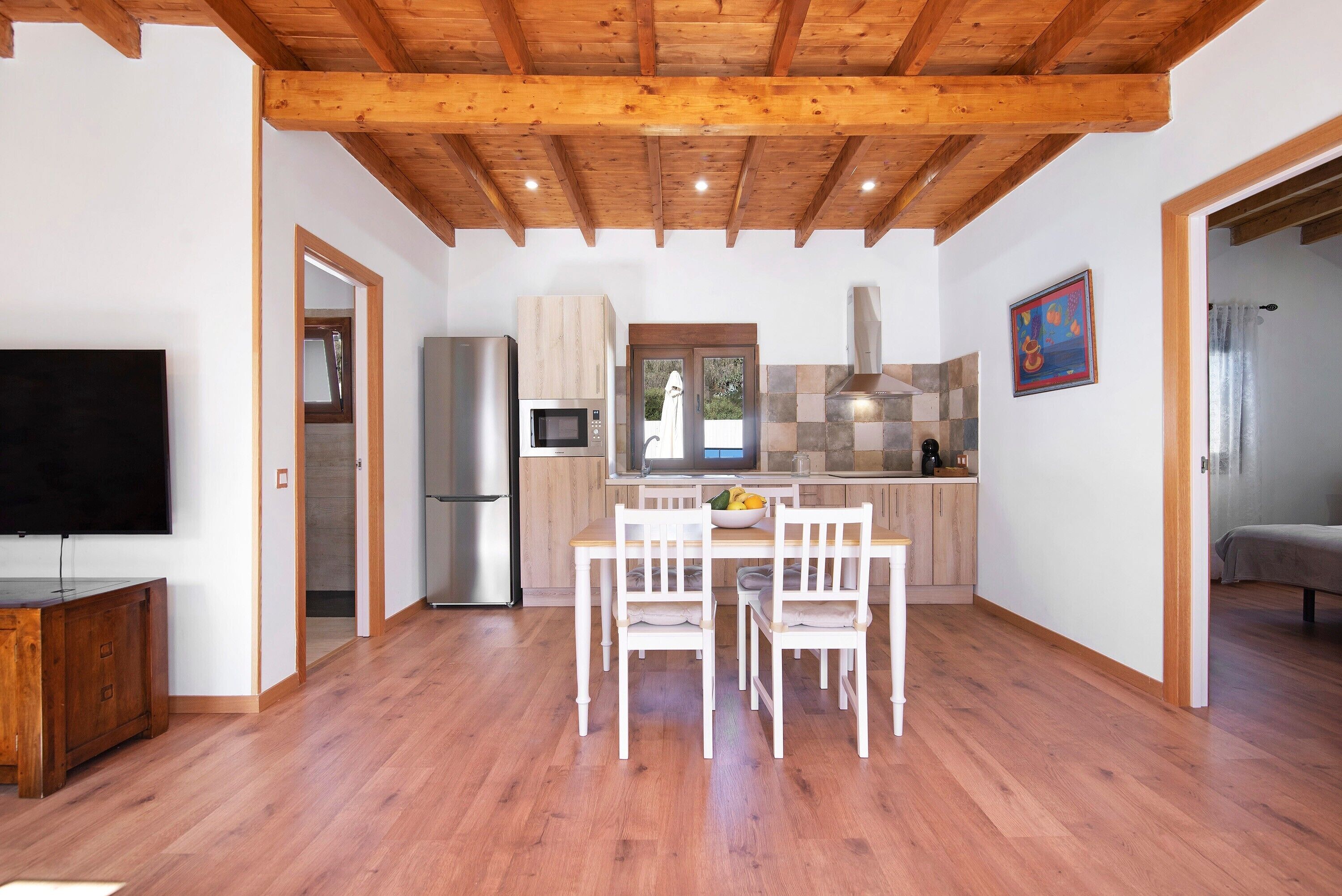 Una cocina moderna con vigas de madera, un comedor con sillas blancas y un televisor de pantalla plana.