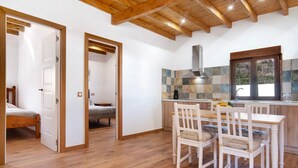 Ein Zimmer mit Holzboden, einem Esstisch mit Stühlen, einer Küchenzeile mit Fliesenrückwand und einem Bett im angrenzenden Raum.