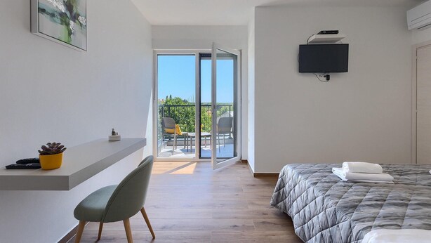A modern hotel room with a balcony, a bed with a grey quilt, a mounted TV, and a small desk with a chair and a potted plant. - Gorgeous home in Pula (Pula)