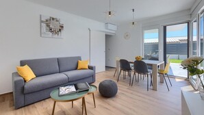 A modern living room with a grey sofa, a small round ottoman, a coffee table, and a dining area with chairs and a table. - Gorgeous home in Pula (Pula)