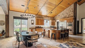 A spacious kitchen with a large island, wooden ceiling, stone wall, and a dining area with chairs. - The Meadowbrook, games, BBQ, huge back yard (Big Bear City)