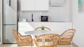 Een moderne keuken met witte kastjes, een ingebouwde magnetron, en een eethoek met rieten stoelen en een kleine potplant.