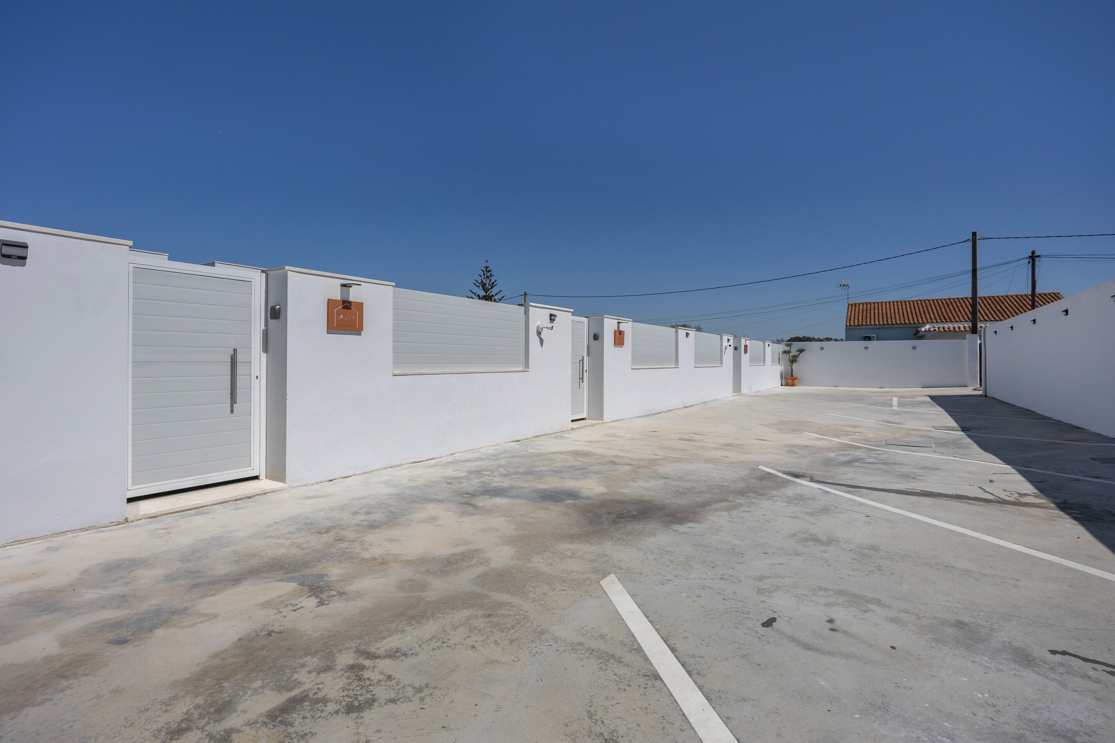 Una hilera de edificios blancos con puertas cerradas, cielo despejado y un área de estacionamiento.