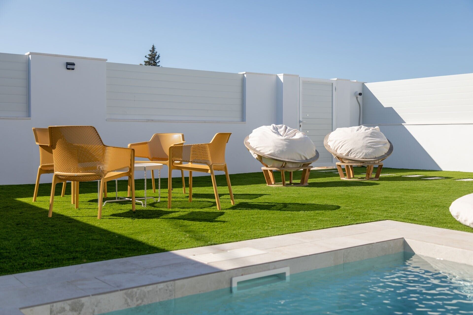 A pool area with a white wall, a small table with chairs, and two bean bags.