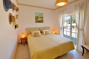 A bedroom with a bed, two bedside lamps, a wall-mounted hat rack, and a window with curtains. - Villa 'Casa Canasta' with Mountain View, Pool and Air Conditioning (São Brás de Alportel)