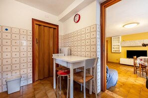 Un coin repas avec une table et des chaises blanches, une horloge au mur et un mur recouvert de carreaux.