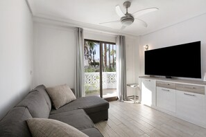 Una sala de estar moderna con un sofá gris, un televisor de pantalla plana y un balcón con vistas a palmeras.