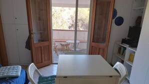 Ein kleines Zimmer mit Bett, Tisch, Stühlen, einem Balkon mit Blick auf Bäume und einem Bücherregal mit Büchern.