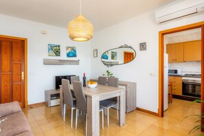 Un espace repas comprenant une table en bois, des chaises, un luminaire suspendu, un téléviseur et une cuisine visible à travers une porte ouverte.