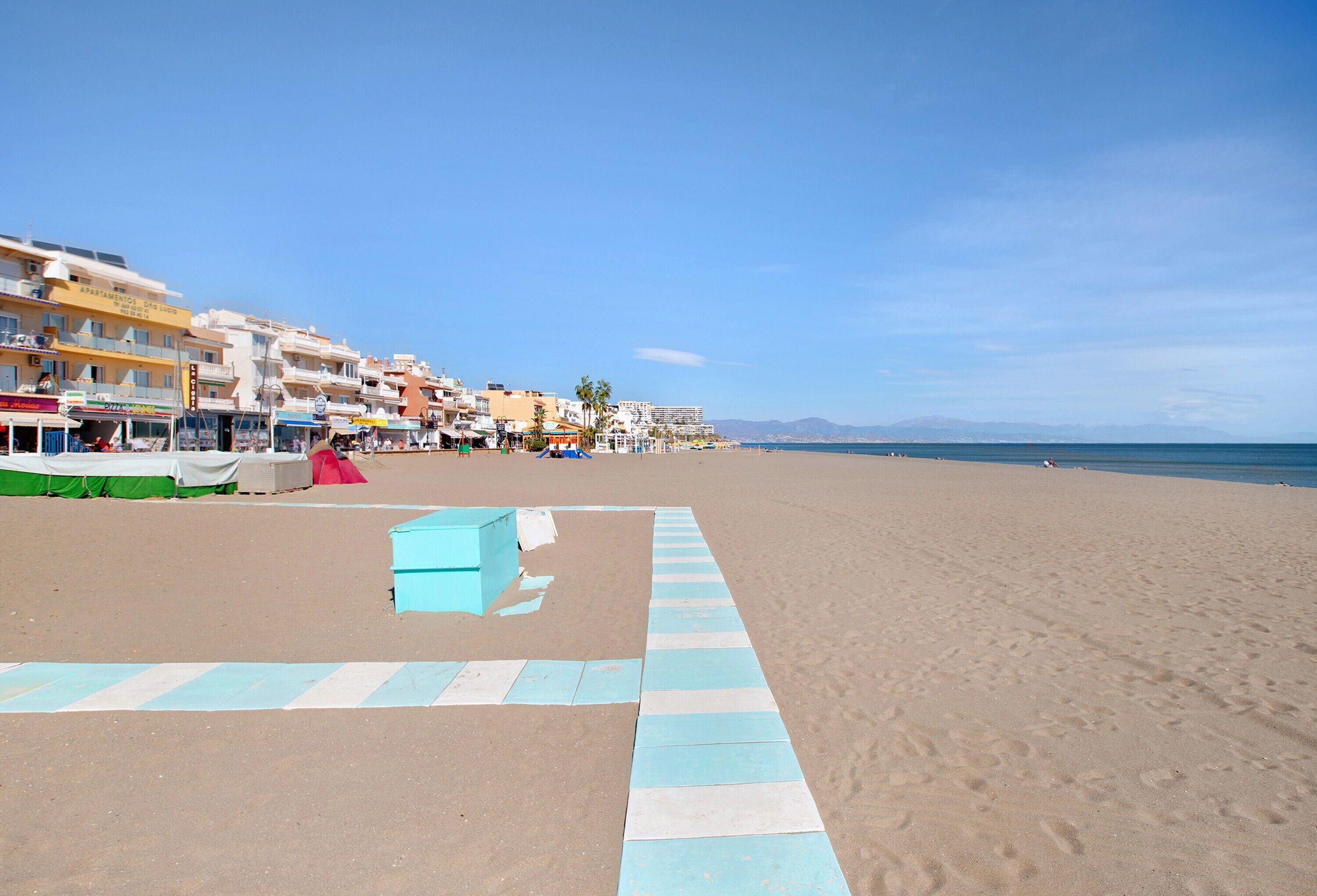 A beach with buildings, a clear sky, and a blue and white striped path.