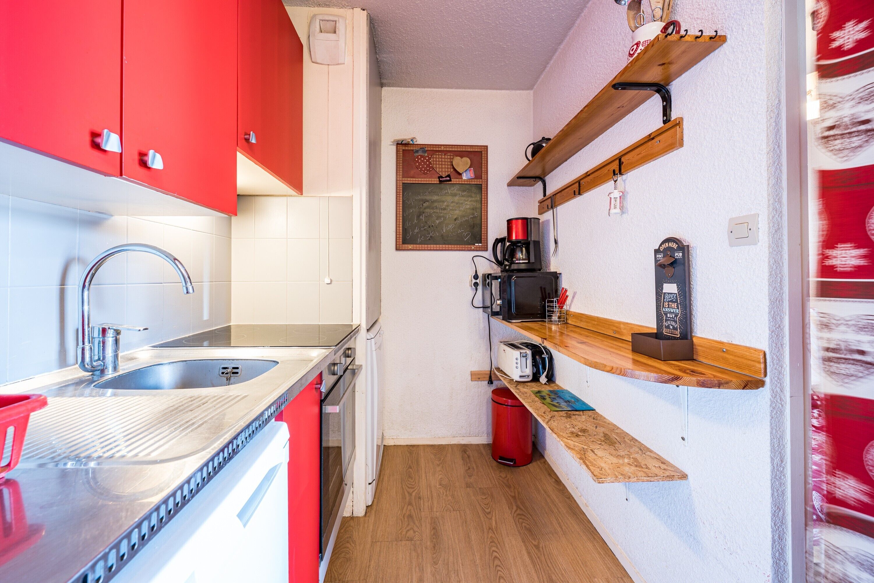 Ett kompakt kök med röda skåp, en diskho i rostfritt stål, en kaffemaskin och hyllor i trä.
