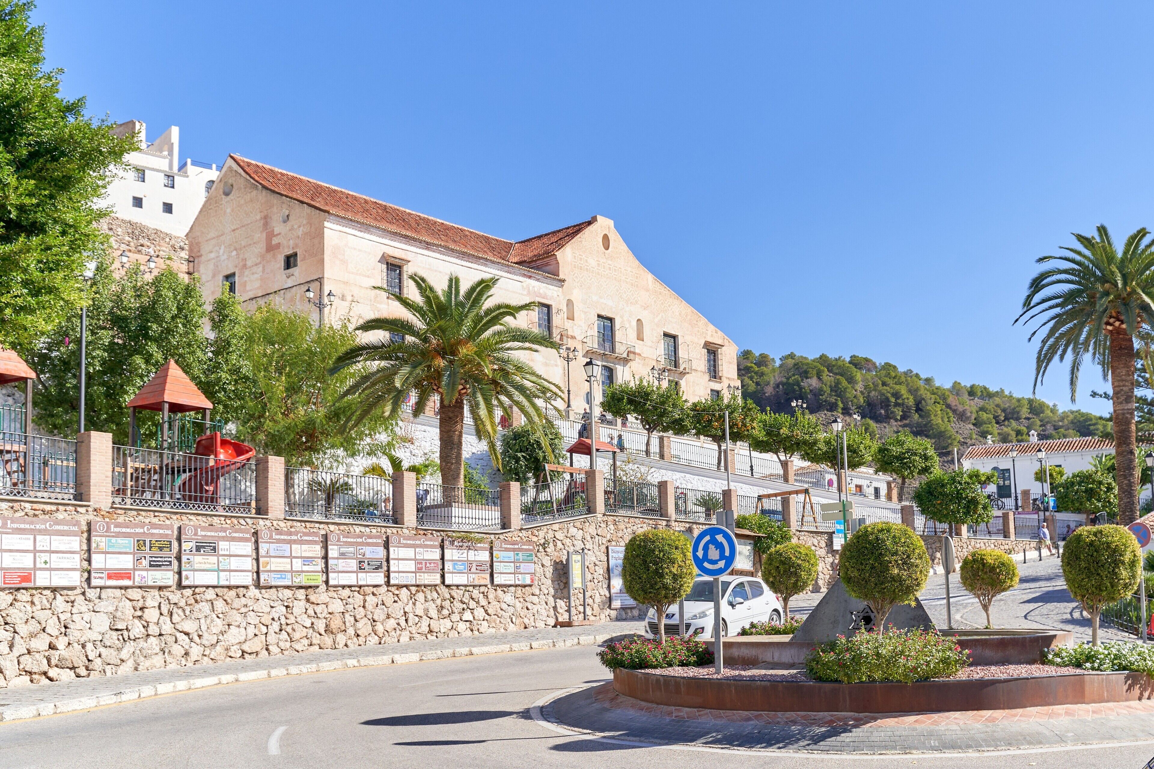 A street with a stone wall, palm trees, and a building with a red roof.