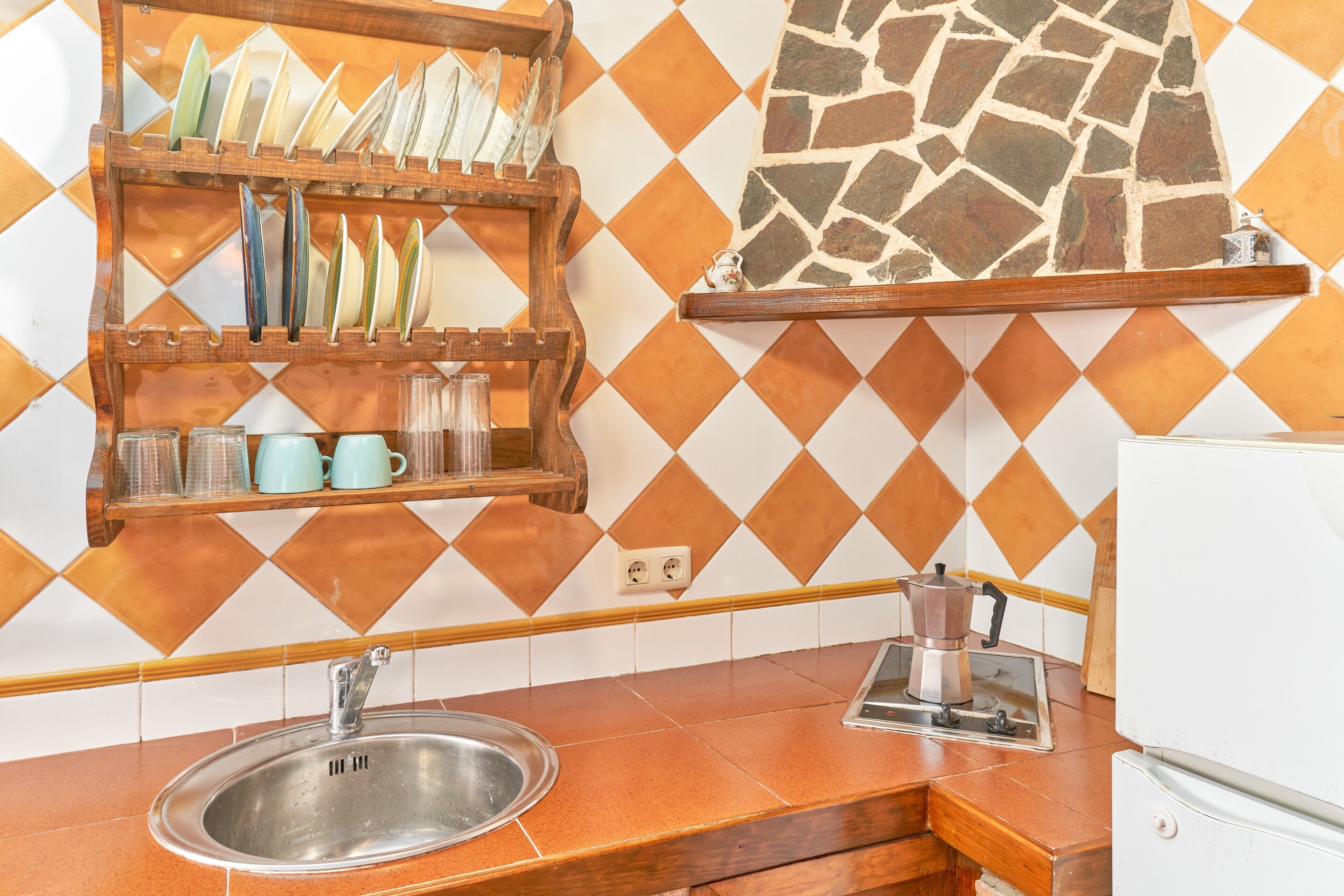 Una cocina con un lavamanos, una cafetera y una alacena con vajilla y tazas.
