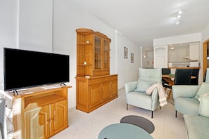 A living room with a flat-screen TV, wooden cabinets, a light blue sofa, and a kitchen area in the background.