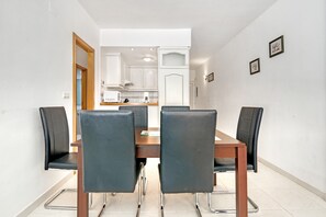 En matplats med ett träbord och fyra stolar, ett kök i bakgrunden och en vit vägg med två inramade tavlor.