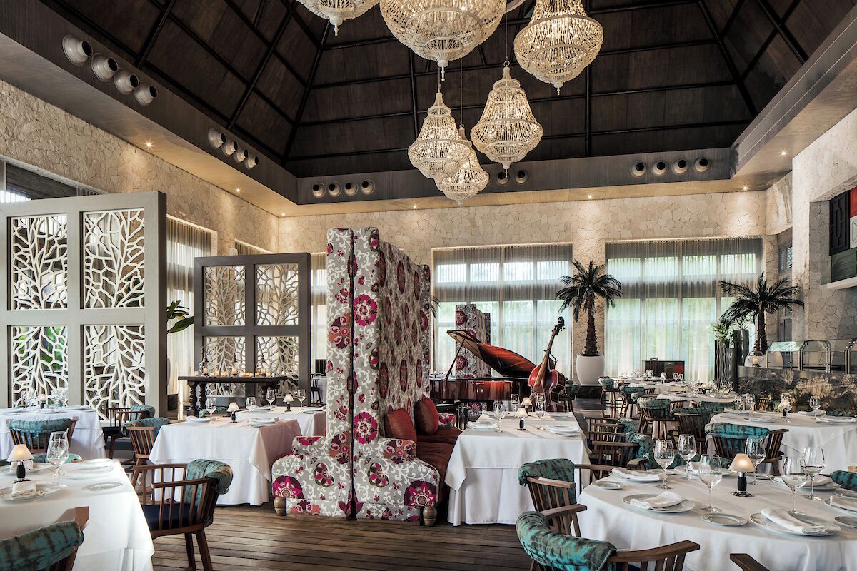 Un comedor espacioso con varias mesas, elegantes lámparas de araña y un amplio ventanal con cortinas.