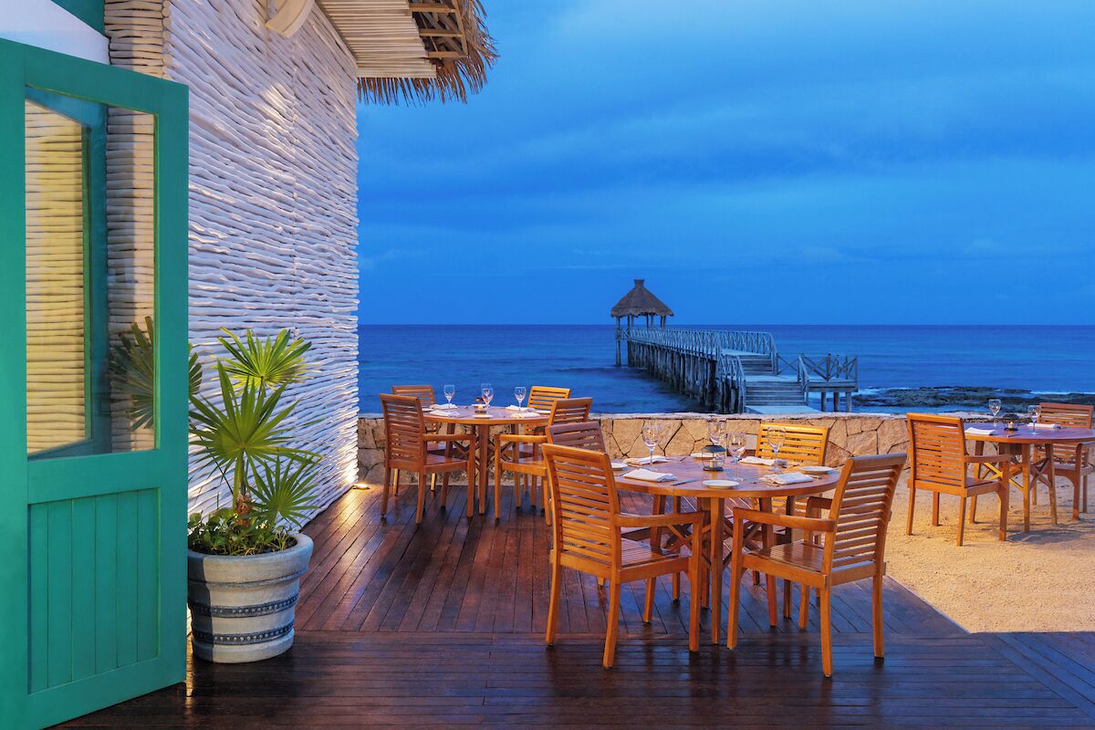 An outdoor dining area with wooden tables and chairs, a potted plant, and a view of the ocean and pier.