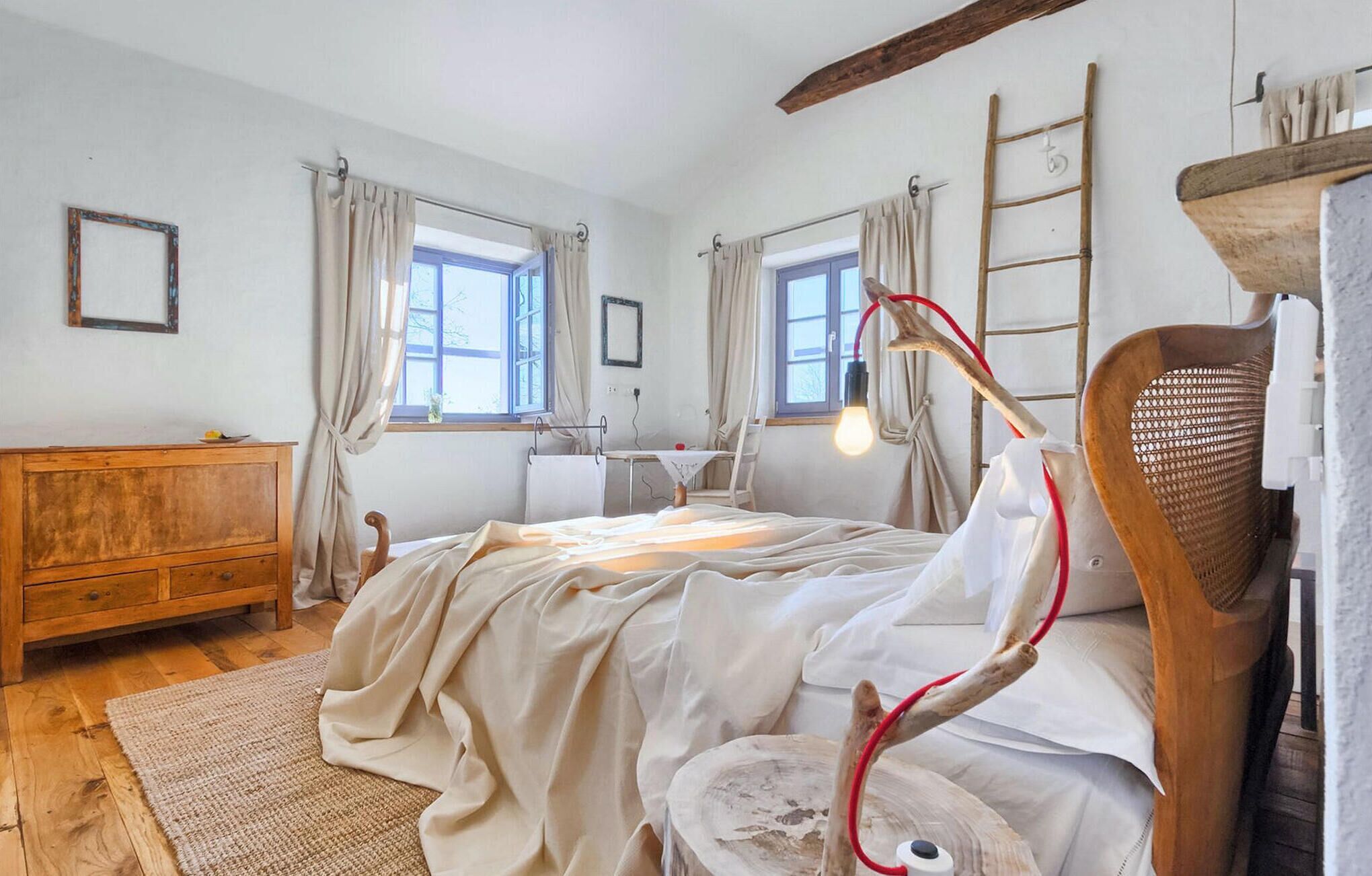 A cozy bedroom with a wooden bed, a wooden dresser, two windows with curtains, a ladder, and a rustic chair.