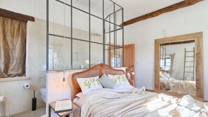 Een slaapkamer met een grote spiegel, een houten plafond, een bed met wit beddengoed, en een nachtkastje met een lamp en een boek.