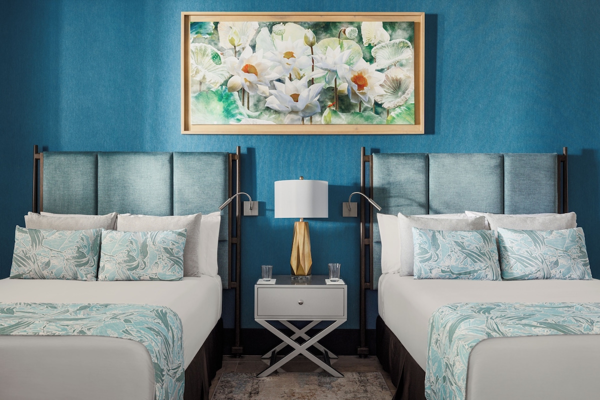 Een hotelkamer met twee bedden, een nachtkastje met een lamp en een bloemistisch kunstwerk in een lijst aan de muur.