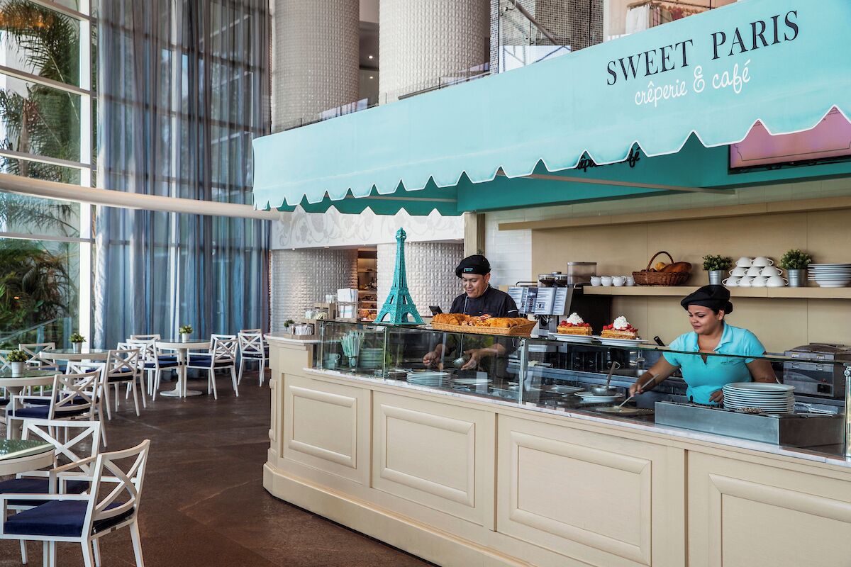 Ein Café namens "Sweet Paris" mit freiem Blick ins Innere, darunter eine Theke, an der ein Koch Speisen zubereitet, ein dekorativer Eiffelturm und ein Sitzbereich mit Tischen und Stühlen.