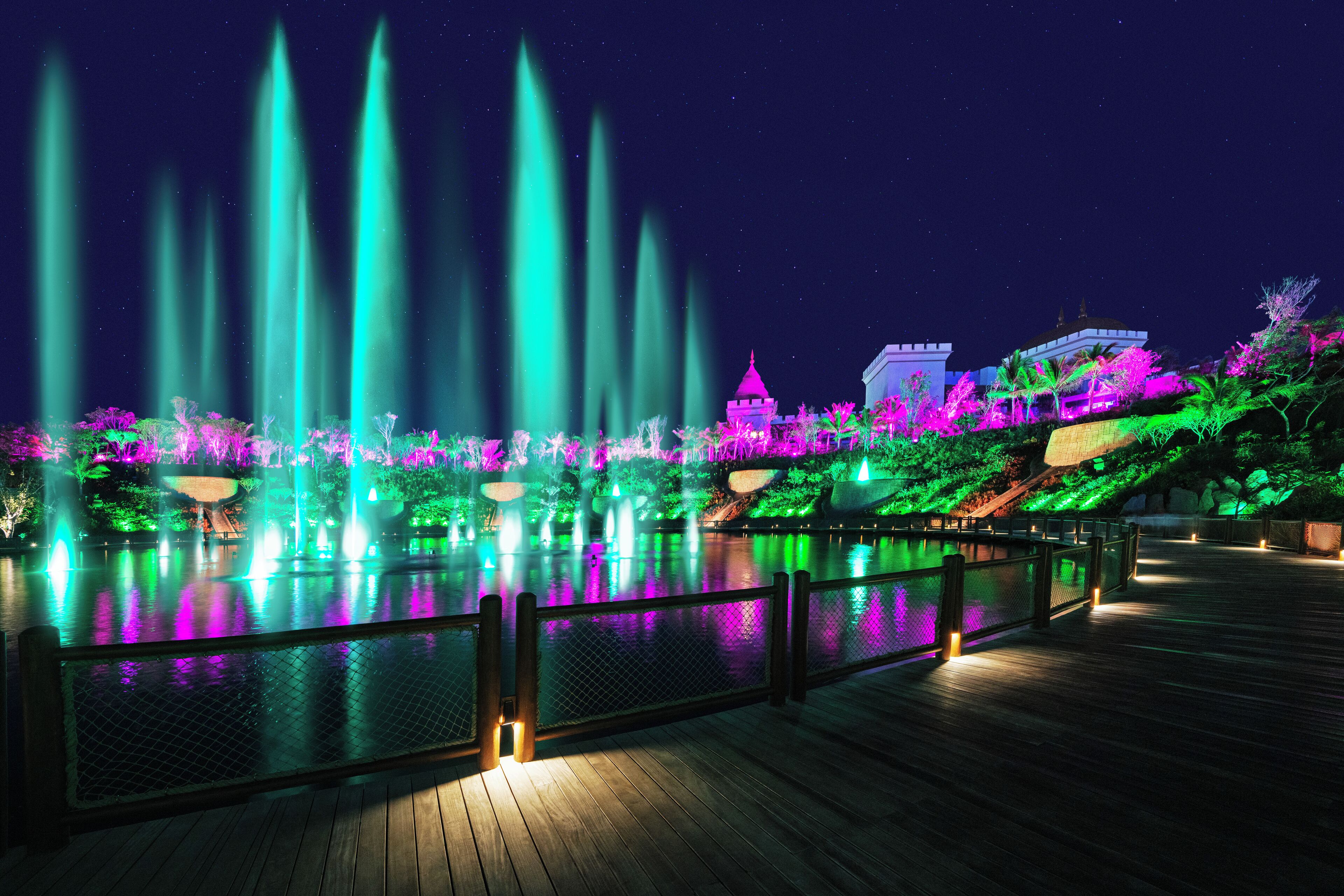 A nighttime view of a water fountain show with illuminated structures and a wooden deck.