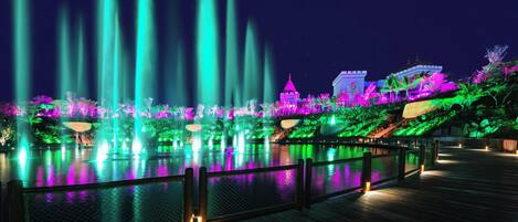 A nighttime view of a water fountain show with illuminated structures and a wooden deck.