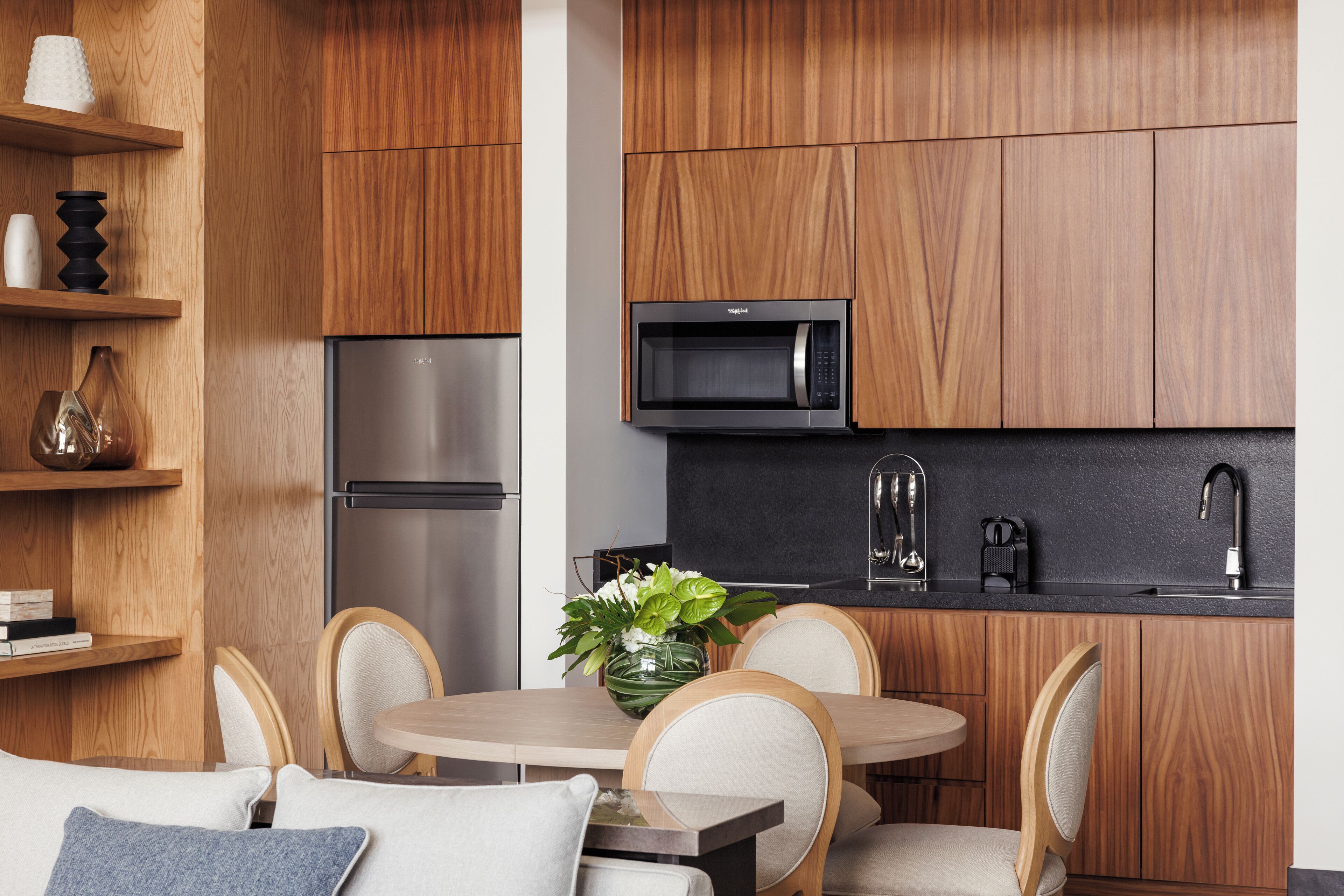 A modern kitchen with wooden cabinets, a stainless steel refrigerator, a microwave, and a sink. There is a dining table with chairs and a vase with flowers on the table.
