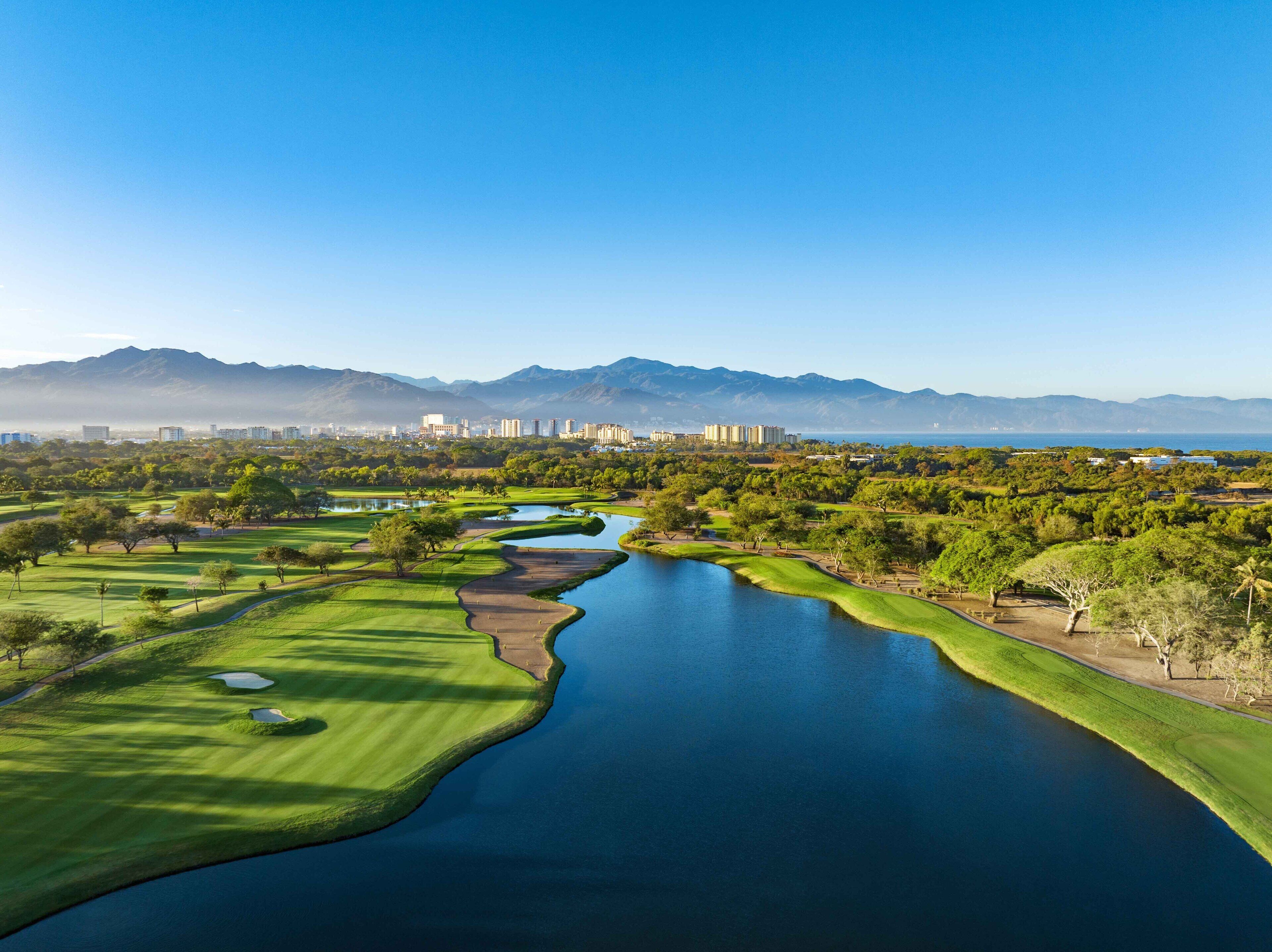 Un campo de golf con un cuerpo de agua, rodeado de vegetación y montañas al fondo.