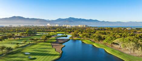 Un campo de golf con un cuerpo de agua, rodeado de vegetación y montañas al fondo.