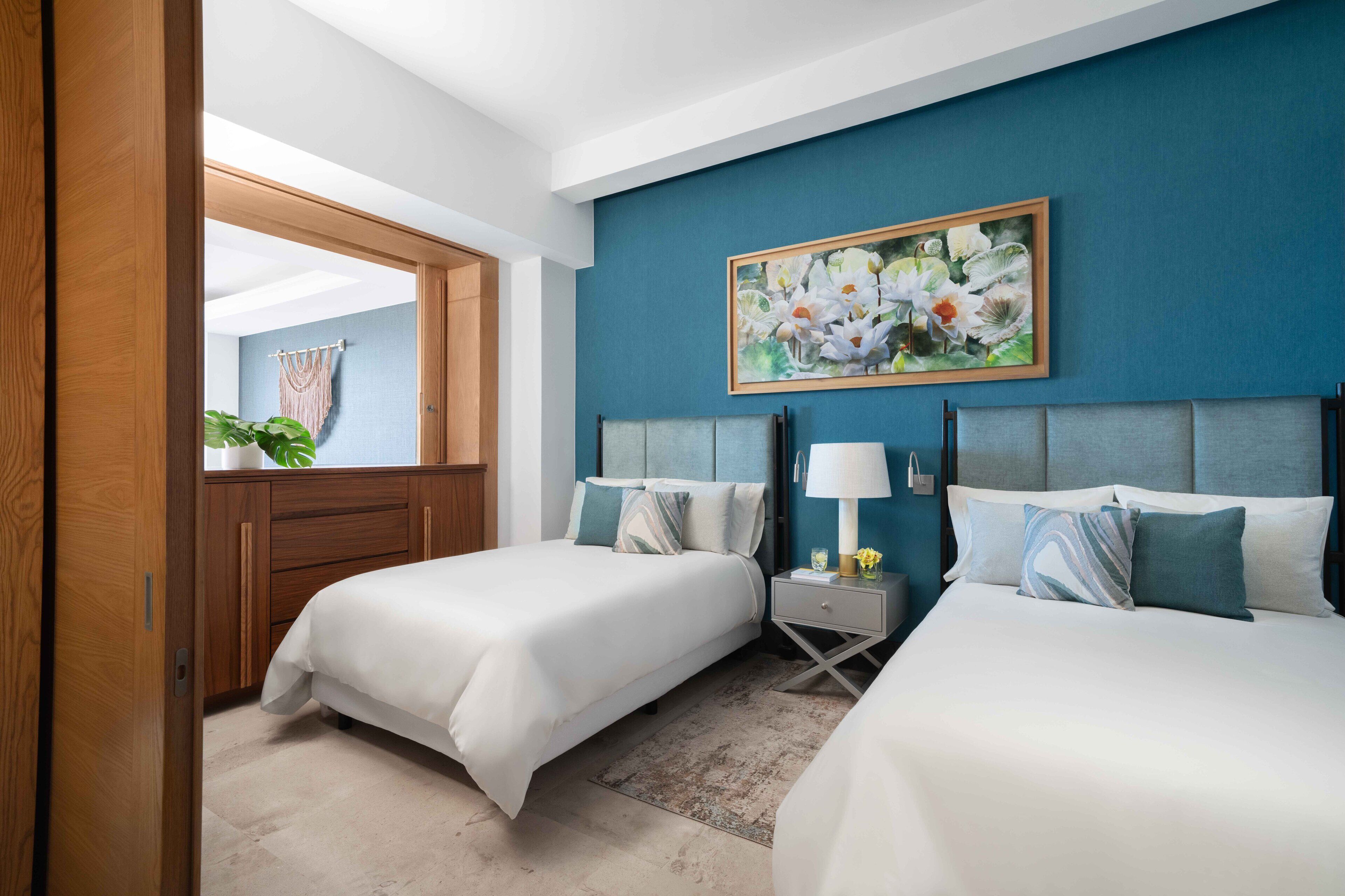 Ein Hotelzimmer mit zwei Betten, einem großen Gemälde an der Wand, einem Nachttisch mit Lampe und einer Holztür.