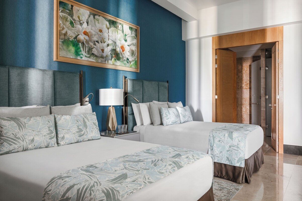 Quarto de hotel com duas camas, um quadro floral grande na parede, cabeceira moderna e porta de madeira que dá para outro cômodo.