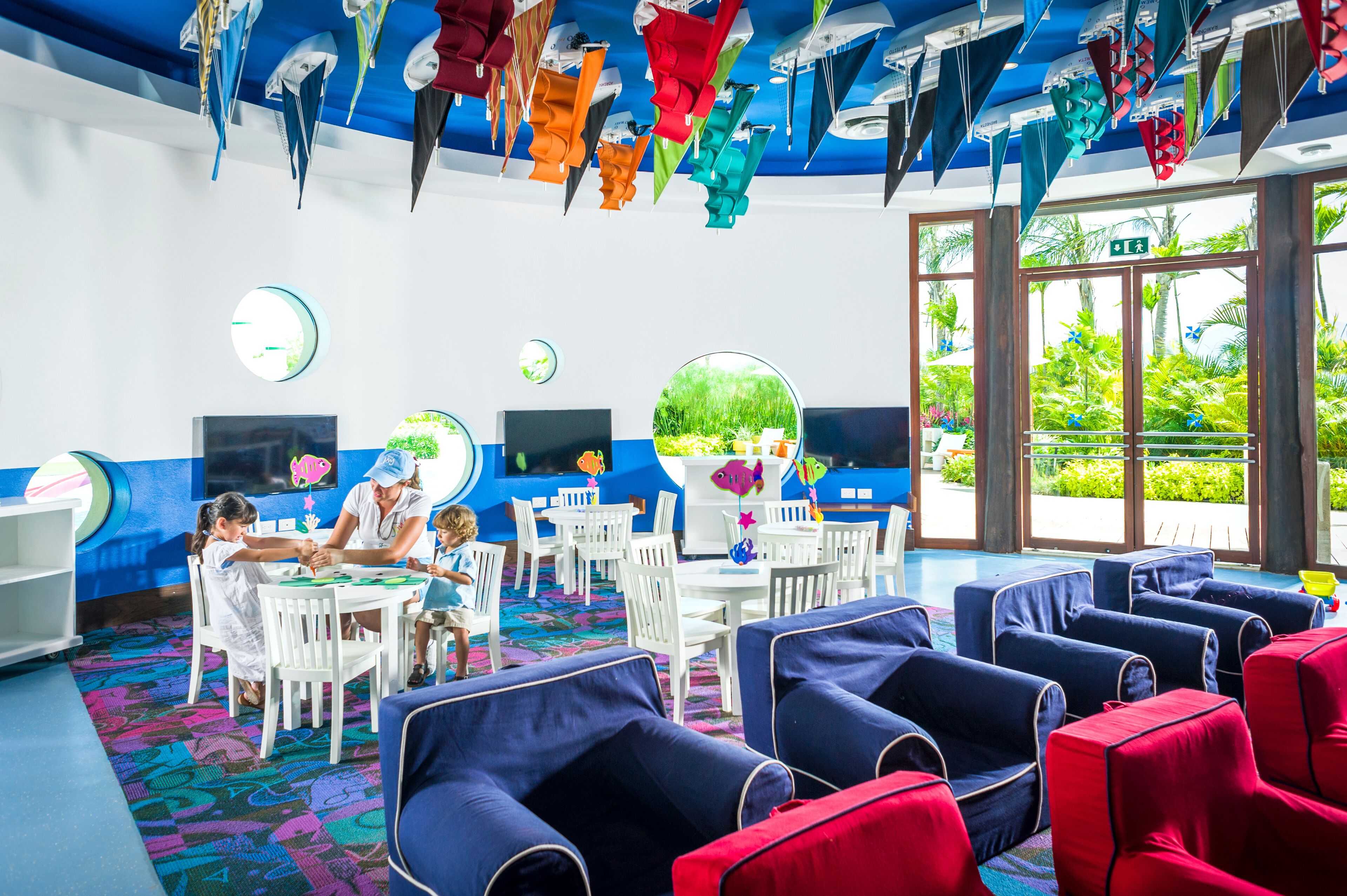 Zona infantil con decoraciones colgantes coloridas, un ventanal con vistas a zonas verdes y áreas de descanso con cojines azules y rojos.