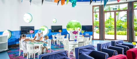 A children's play area with colorful hanging decorations, a large window with a view of greenery, and seating areas with blue and red cushions.