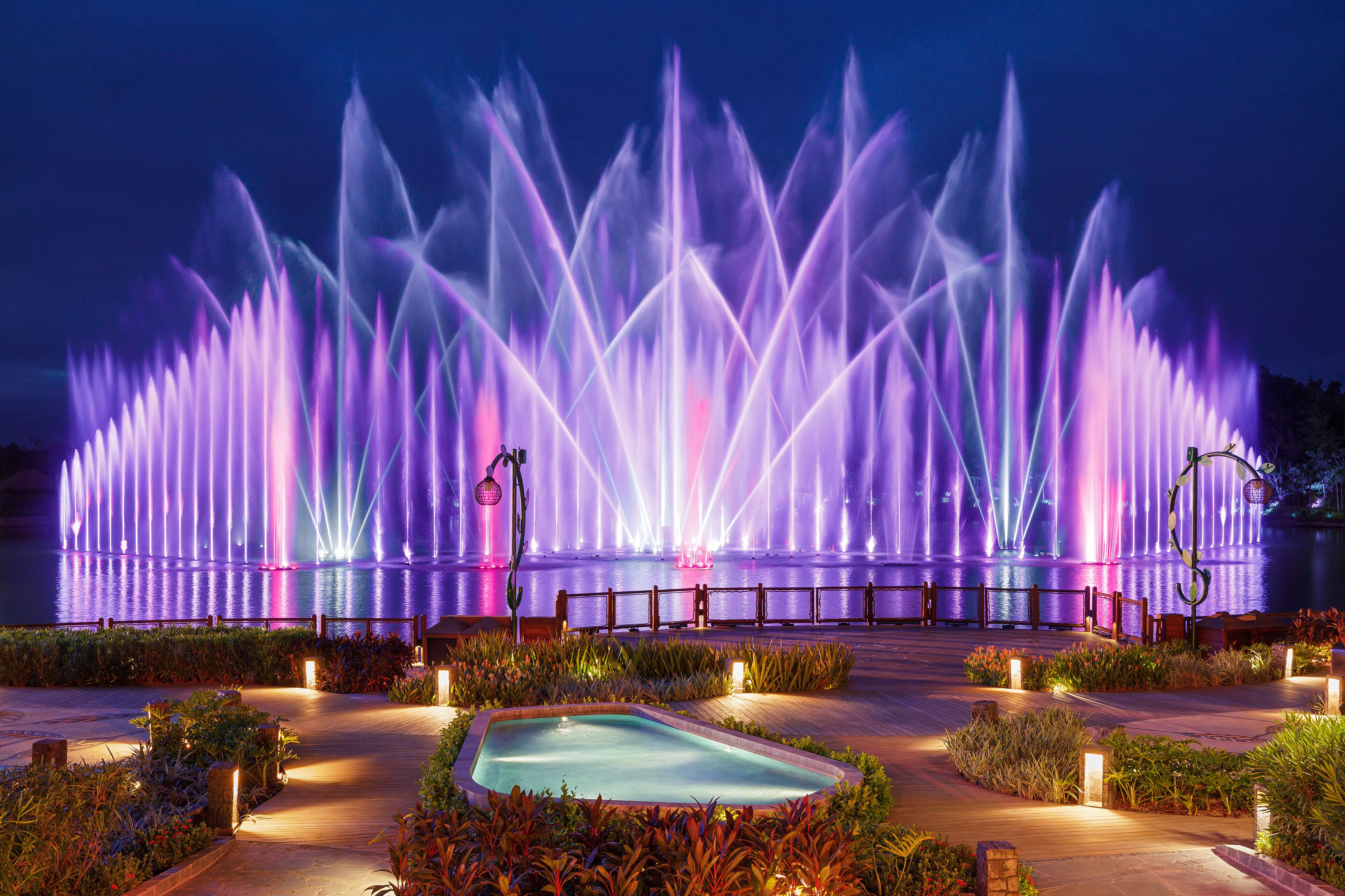A large outdoor fountain with water jets illuminated in purple and pink lights, surrounded by a landscaped garden with a small pool and benches.