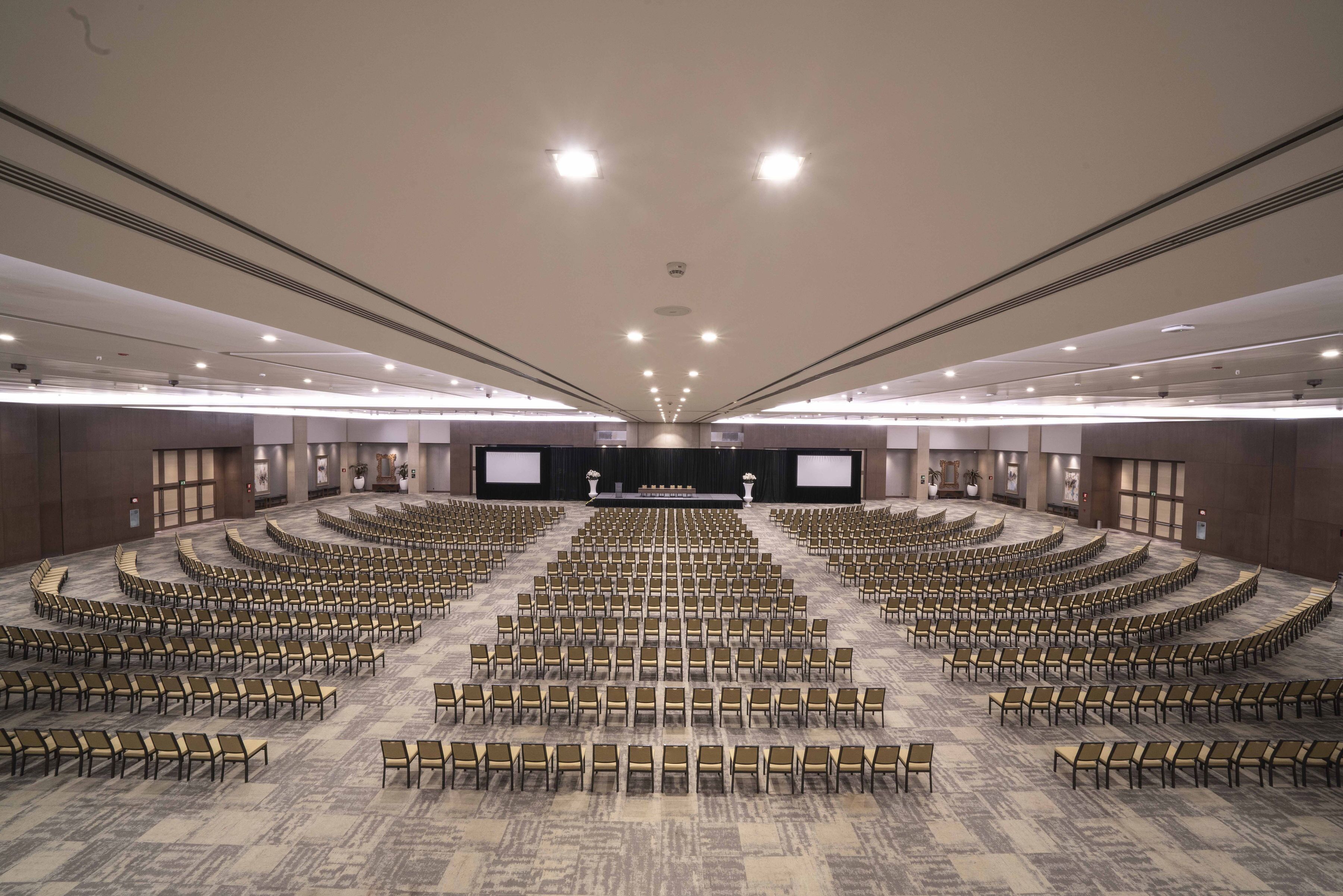 A large conference room with rows of chairs arranged for a presentation, a stage with a screen, and a carpeted floor.