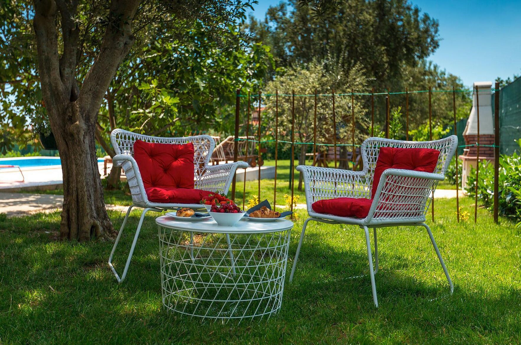 Two white wicker chairs with red cushions, a round metal table, and a bowl of fruit and pastries.