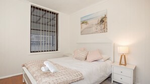 Une chambre à coucher comprenant un lit, une table de chevet, une lampe, une fenêtre avec des stores et un tableau sur le thème de la plage accroché au mur.