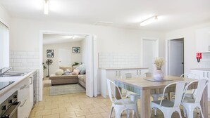 Une cuisine moderne avec des meubles blancs, un coin repas avec une table en bois et des chaises blanches, et un espace de vie chaleureux avec un canapé et des plantes.