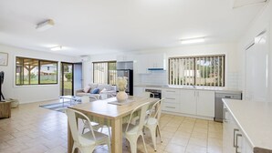 Une cuisine moderne avec des meubles blancs, un coin repas doté d’une table en bois et de chaises blanches, et une grande fenêtre avec des stores.