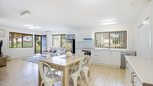 A modern kitchen with white cabinets, a dining area with a wooden table and white chairs, and a large window with blinds.
