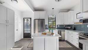 A modern kitchen with white cabinets, a stainless steel refrigerator, a built-in oven, and a large island with a bowl of popcorn and bottles. - Pet-Friendly +Historic Charm & Modern Comfort Near Upper King (Charleston)