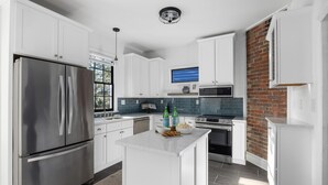 A modern kitchen with white cabinetry, stainless steel appliances, a central island, and a brick accent wall. - Pet-Friendly +Historic Charm & Modern Comfort Near Upper King (Charleston)