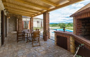 Une terrasse couverte avec une cheminée en briques, une piscine et une vue sur la campagne.