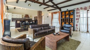 Un vaste espace de vie comprenant un canapé en cuir foncé, une table basse en bois et une bibliothèque. On aperçoit à l’arrière-plan une cuisine avec un mur en briques et des meubles blancs.