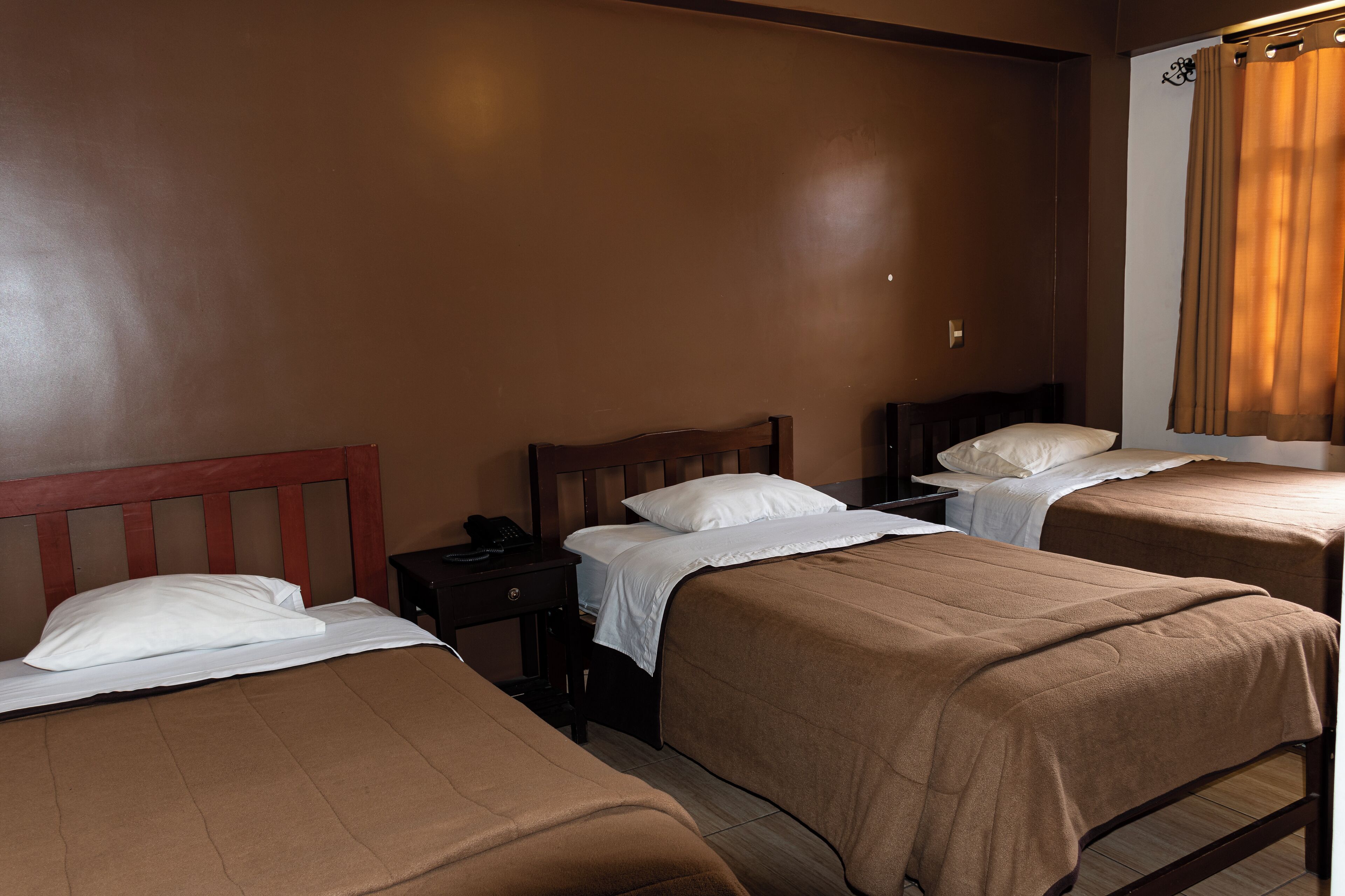 A hotel room with three single beds, each with a wooden headboard and white bedding. The walls are painted brown, and there is a window with a curtain.