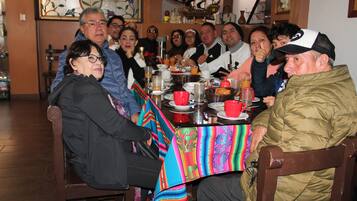 Un grupo de personas sentadas alrededor de una mesa con diversos platos y bebidas, en una habitación con muebles de madera y decoración en la pared.